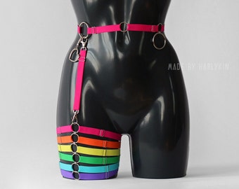 May include: A rainbow-colored thigh harness with silver rings and a pink strap attached to a silver heart-shaped charm. The harness is worn on a black mannequin.
