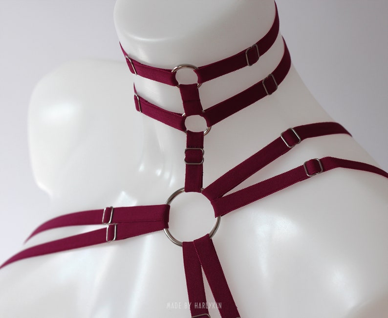 May include: Burgundy colored harness with multiple straps and silver metal rings. The harness has a choker style neckline with two straps and a ring.  The straps are adjustable with silver metal buckles.  The harness is made by Hardykin.