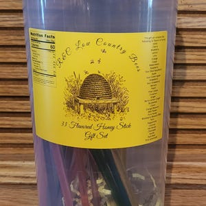 May include: A clear plastic container with a blue lid, filled with flavored honey sticks. A yellow label reads "R&C Low Country Bees" and "33 Flavored Honey Stick Gift Set". The container includes a variety of colorful honey sticks.