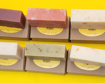 May include: An assortment of handmade soap bars in various colors, including beige, burgundy, and green, displayed on individual cardboard boxes with yellow labels. The soaps are arranged on a bright yellow surface.