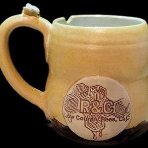 May include: A handmade ceramic mug with a yellow glaze. The handle has a small bee figurine attached. A white circular emblem with a honeycomb design and the text "R&C Low Country Bees, LLC" is on the side.