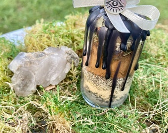 May include: A small glass bottle with a black wax seal and a white ribbon tied around the neck. The bottle contains a mixture of herbs and crystals. A silver charm with an eye symbol hangs from the ribbon.