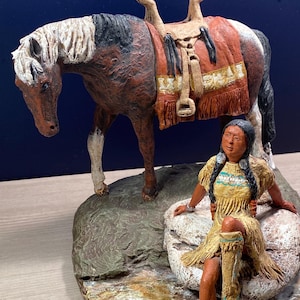 May include: A detailed figurine depicting a Native American woman seated near a horse. The horse has a saddle and is brown, white, and black. The woman wears a yellow dress and sits on a rock. The base is a textured, oval shape.