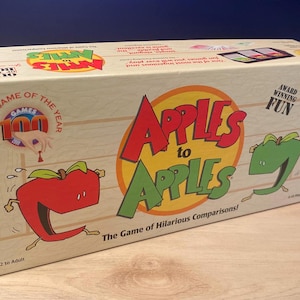 Puede incluir: Caja de color claro para el juego "Apples to Apples" con texto rojo y verde e ilustraciones de manzanas. La caja tiene el texto "Party Game of the Year" y "Award Winning Fun". El juego es para mayores de 12 años.