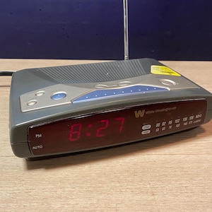 Peut inclure: Un radio-réveil numérique gris White-Westinghouse avec un affichage LED rouge indiquant 8:27. Il possède une antenne argentée et des accents bleus. L'horloge affiche les fréquences radio FM et AM.
