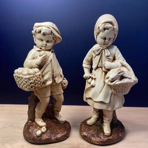 May include: Two antique bisque figurines of children. The boy wears a cap and carries a basket of fruit. The girl wears a bonnet and carries a basket of fruit. Both figures are in a cream color and stand on brown bases.