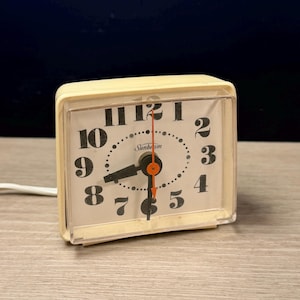 May include: A vintage cream-colored Sunbeam alarm clock with a square face. The clock has black numerals, black hands, and an orange second hand. The clock face is protected by a clear plastic cover.