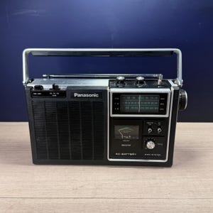 Könnte beinhalten: Ein schwarzes und silbernes Panasonic-Tragradio mit oberem Tragegriff, Teleskopantenne, Frequenzanzeige, Signalstärkeanzeige und Lautsprechergitter. Auf der Vorderseite befinden sich die Aufschriften Panasonic und AC-BATTERY.