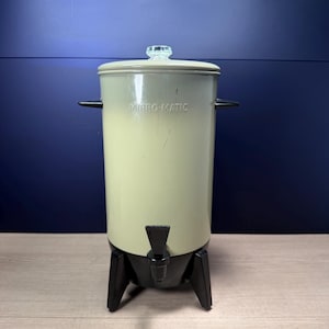 May include: A vintage Mirro-Matic beverage dispenser with a cylindrical cream-colored body, a black base with three legs, and a black spigot. The lid features a clear glass knob handle. The brand name Mirro-Matic is embossed on the front surface.