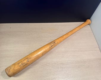 May include: A light brown wooden baseball bat with a smooth finish. The barrel features the black printed text Louisville Slugger and Genuine, along with a signature and a circular logo stamp containing the text Louisville Slugger.