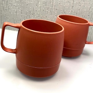 May include: Two terracotta colored ceramic mugs with handles. The mugs have a rounded shape and a smooth finish.