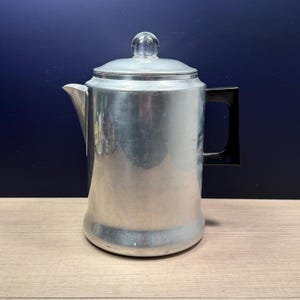 May include: A vintage aluminum coffee percolator with a clear glass knob on the lid and a black handle. The coffee pot has a spout for pouring and is designed for stovetop use. The metal has a brushed finish.
