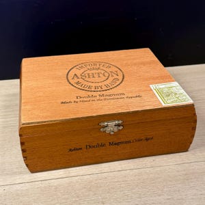 May include: A rectangular wooden cigar box with a light brown finish and a metal latch. The lid features the text Imported Ashton Made By Hand Double Magnum Made by Hand in the Dominican Republic, with a green tax stamp and side text reading Ashton Double Magnum Cedar Aged.