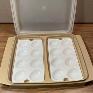 May include: A beige Tupperware egg tray container with a translucent lid. The base features two removable white plastic inserts, each containing eight oval-shaped indentations designed to hold sixteen eggs in total for storage and transport.
