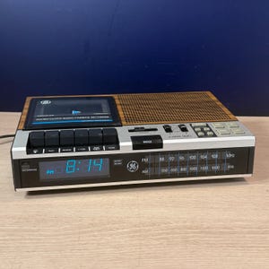 Op de afbeelding: Een GE FM/AM klokradio met cassetterecorder, voorzien van een houtlook bovenkant, een digitaal display met 8:14 PM en een zilverkleurig bedieningspaneel. Het apparaat heeft zwarte afspeelknoppen, een volumeschuifregelaar, een sluimerknop en een tuner.