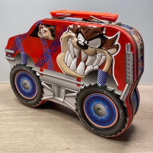May include: Red metal lunchbox featuring a cartoon Tasmanian Devil monster truck. The truck has a red body, black tires, and a blue and white design. The Tasmanian Devil is grinning with sharp teeth and is wearing a blue and white striped shirt.