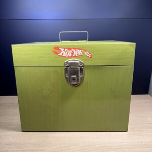 Op de afbeelding: Een groene houten kist met een metalen sluiting en handvat. De kist heeft een rood en wit Hot Wheels logo sticker. De rechthoekige kist heeft een vintage uitstraling.