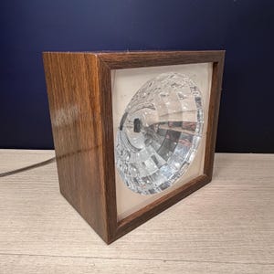 May include: A square table lamp featuring a dark brown wood-grain frame and a central, circular, faceted clear glass lens. The lamp includes a visible internal light bulb filament and a thin electrical cord extending from the side of the frame.