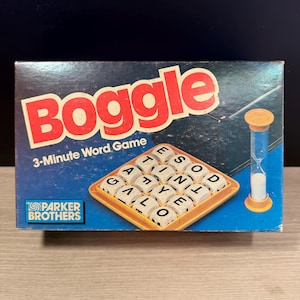 Pode incluir: Caixa de jogo de palavras Boggle vintage com a palavra "Boggle" em letras vermelhas grandes. A caixa azul apresenta as palavras "3-Minute Word Game" e o logotipo da Parker Brothers. O jogo inclui uma grelha de dados com letras e um cronômetro de 3 minutos.