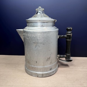 May include: Vintage aluminum coffee pot with a glass lid and a black and wood handle. The pot has a spout and a decorative band around the top. The coffee pot is sitting on a wooden surface.