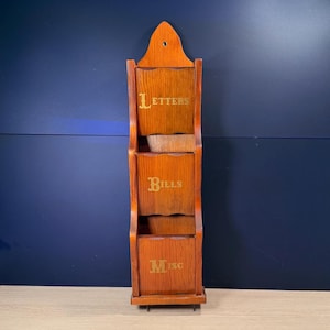 May include: A wooden wall-mounted organizer with three compartments labeled "Letters", "Bills", and "Misc" in gold lettering. The organizer is made of stained wood and has a decorative top with a hole for hanging.