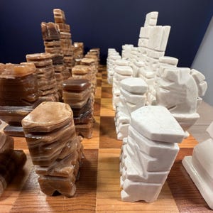 May include: A chess set with pieces carved from stone. The pieces are in shades of brown and white, with a layered, textured appearance. The chessboard is made of wood with a classic checkerboard pattern.