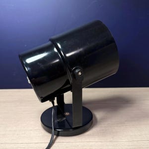 May include: A black spotlight with a cylindrical head and a circular base. The spotlight is angled upwards and has a black stand. A power cord extends from the base. The background is a gradient of blue.