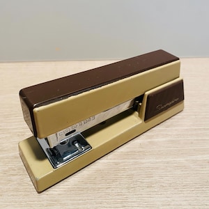 May include: A brown and beige metal stapler with a silver staple remover. The stapler has a label that says "Lift Cap to Load". The stapler is made by Swingline.