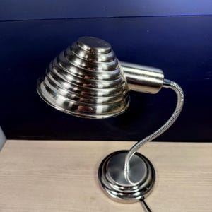 May include: A silver-colored desk lamp with a conical, ridged shade. The lamp has a flexible, gooseneck arm and a circular base. The lamp is made of metal and has a modern design.