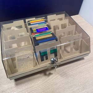 May include: A vintage beige and clear plastic floppy disk storage case with a lock and key. The case is filled with colorful 3.5-inch floppy disks and dividers. Some handwritten notes are visible inside the case.