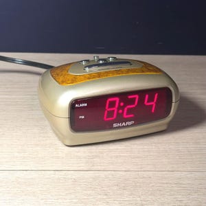 May include: A Sharp brand digital alarm clock with a champagne-colored plastic casing and a faux wood grain top panel. The front features a dark red display showing the time 8:24 in bright red digital numerals, with labels for ALARM and PM.