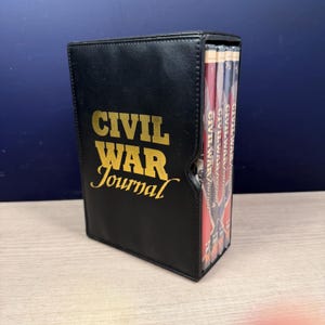 Może przedstawiać: Czarny skórzany futerał na dziennik ze złotymi literami z napisem "CIVIL WAR Journal". Etui zawiera kilka książek z tytułem "CIVIL WAR Journal" widocznym na grzbietach. Książki mają czerwone i białe okładki.