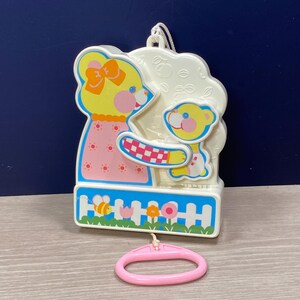 May include: A vintage baby mobile featuring a yellow bear with a pink bow and dress, holding a checkered object, and a smaller bear. The mobile has a white background with a blue and white fence design, a bee, and flowers. A pink oval-shaped pull is attached.