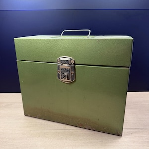 Pode incluir: Uma caixa de armazenamento de metal verde vintage com uma alça e um fecho de trava. A caixa tem formato retangular e aparência ligeiramente desgastada, sugerindo que foi usada. A alça é de metal e é fixada na parte superior da caixa.
