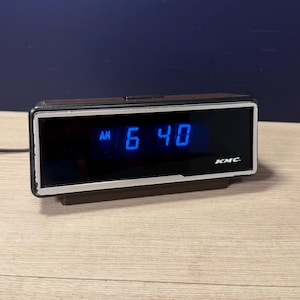 Pode incluir: Um relógio digital vintage exibindo "AM 6:40" em algarismos azuis. O relógio tem um mostrador preto, uma moldura prateada e uma parte superior marrom. A marca "KMC" é visível. O relógio está sobre uma superfície de madeira.