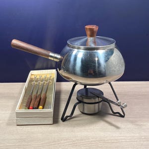 Puede incluir: Un juego de fondue vintage con una olla de acero inoxidable, mango y tapa de madera. El juego incluye un soporte, un quemador y una caja de tenedores de fondue con puntas de colores. La olla tiene aproximadamente 15 cm de diámetro.