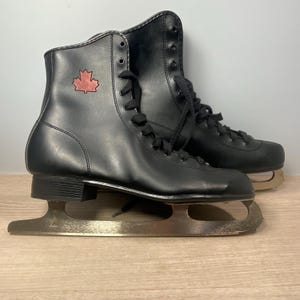 May include: Pair of black ice skates with a red maple leaf emblem on the side. The skates have black laces and silver blades. The skates are on a wooden surface.