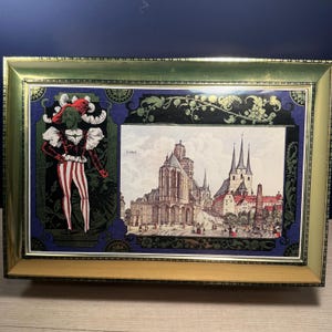 May include: A framed print with a gold-coloured frame. The print features a colourful illustration of a jester on the left and a detailed depiction of a European city with tall spires on the right. The word "Erfurt" is visible.