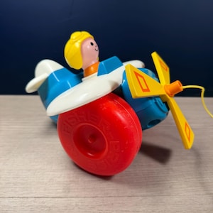 Könnte beinhalten: Ein Vintage-Spielzeugflugzeug von Fisher-Price in Blau, Weiß, Rot und Gelb. Das Spielzeug hat einen lächelnden Piloten mit gelbem Haar. Das rote Rad hat das Fisher-Price-Logo. Der Propeller ist gelb mit orangefarbenen Akzenten. Ein gelber Faden ist befestigt.