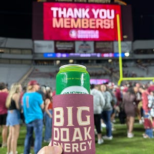 May include: A maroon can cooler with the text "BIG OAK ENERGY" in gold lettering. A green can of a beverage is inside the cooler.