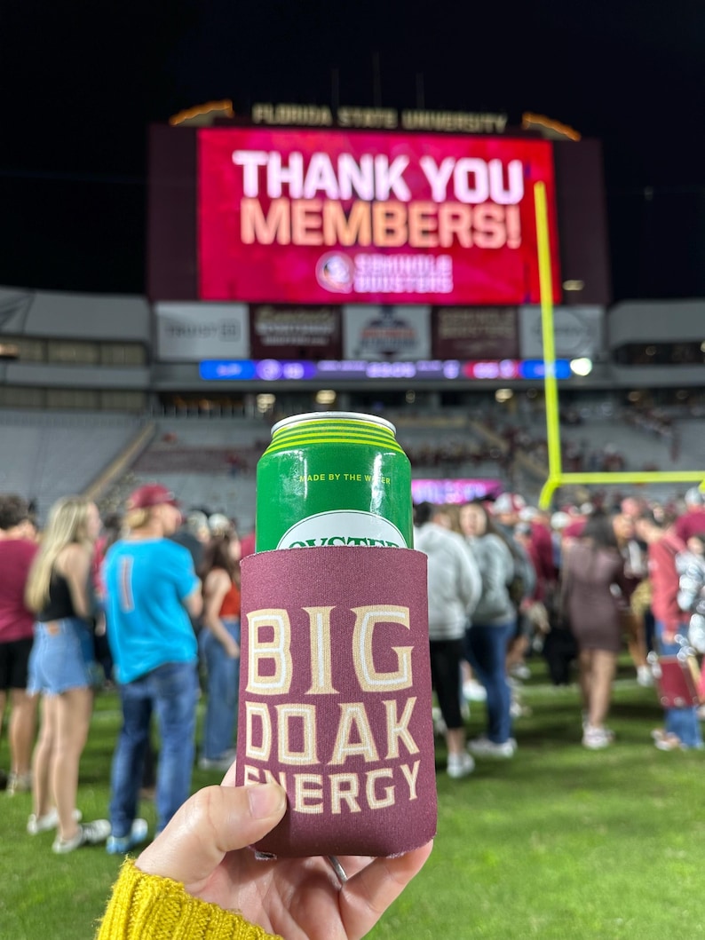 May include: A maroon can cooler with the text "BIG OAK ENERGY" in gold lettering. A green can of a beverage is inside the cooler.