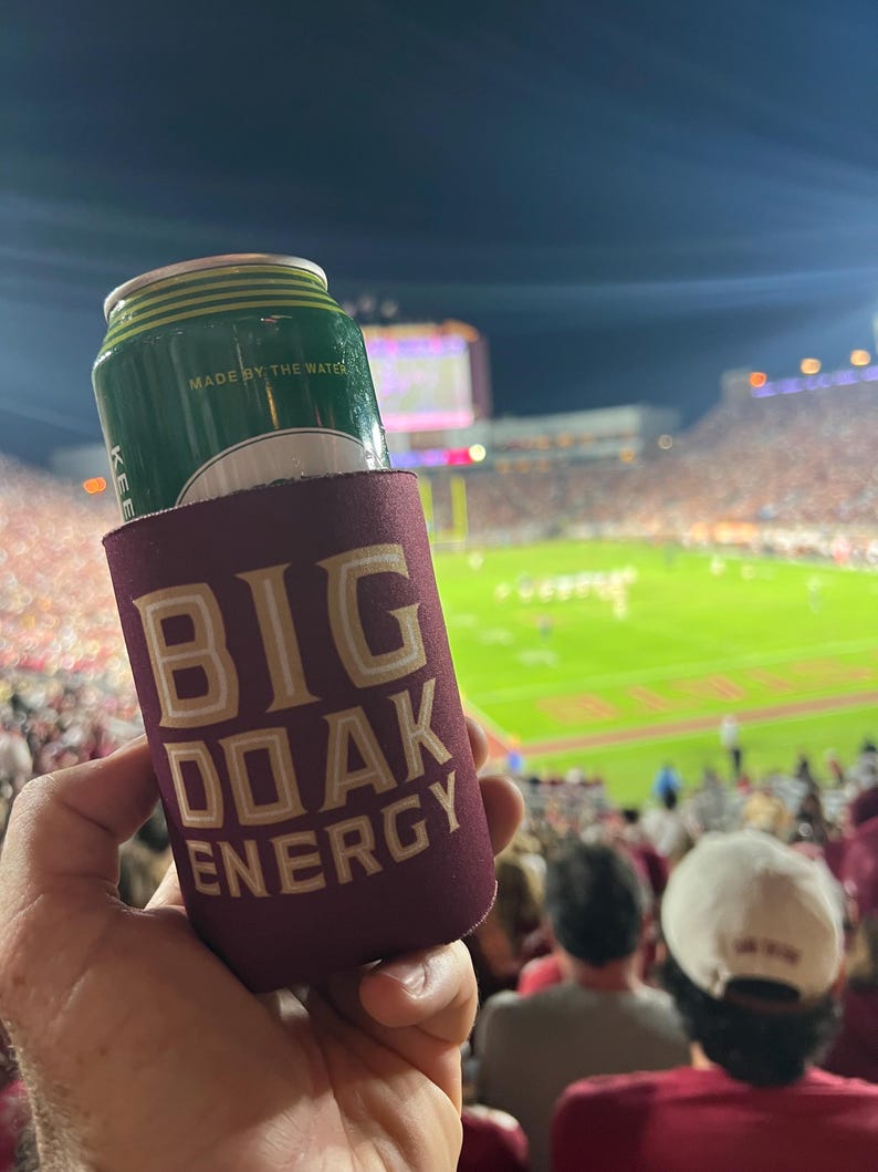 May include: A maroon can cooler with the text "BIG OAK ENERGY" in gold lettering. A green can with the text "MADE BY THE WATER" is inside the cooler.