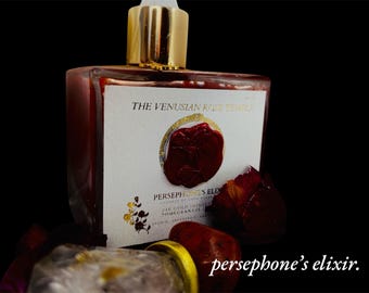 Può includere: Una bottiglia di vetro con un tappo dorato e un liquido rosso all'interno. L'etichetta recita "The Venusian Rose Temple Persephone's Elixir 24k Gold Infused Pomegranate Oil".