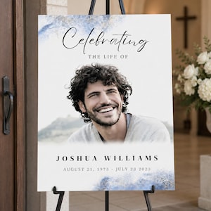 May include: A memorial sign on a black easel. The sign features a photo of a smiling man with the text "Celebrating The Life Of" above the photo. Below the photo is the name "JOSHUA WILLIAMS" and dates "AUGUST 21, 1975 - JULY 23 2023".