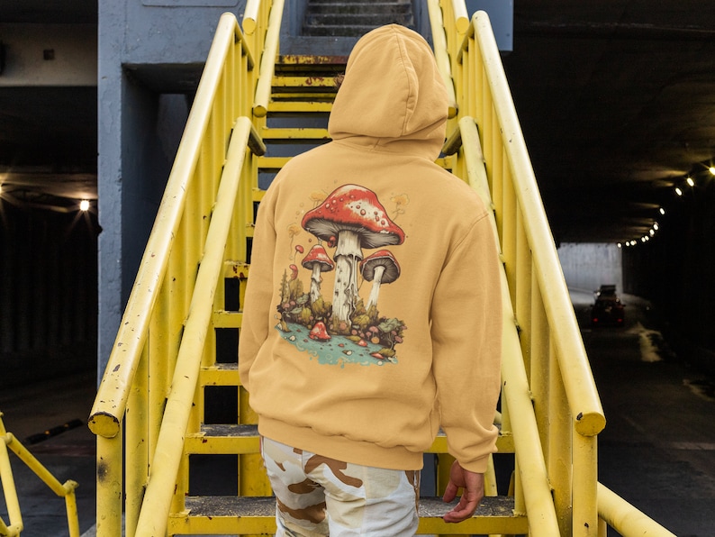 May include: A mustard yellow hoodie with a graphic of red and white mushrooms on the back. The hoodie has a drawstring hood and a kangaroo pocket.