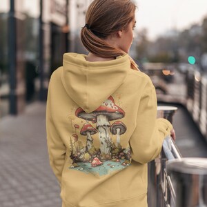 May include: A yellow hoodie with a graphic of red and white mushrooms in a forest setting. The mushrooms are surrounded by green leaves and a blue stream.