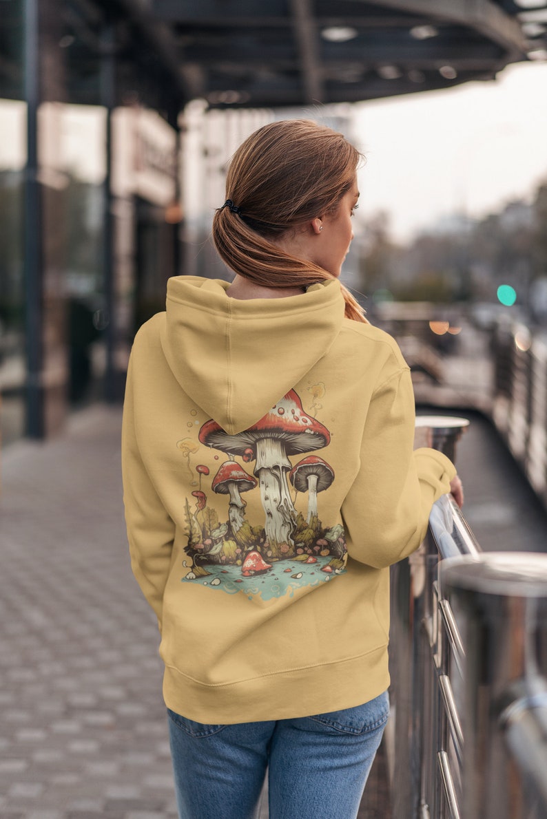 May include: A yellow hoodie with a graphic of red and white mushrooms in a forest setting. The mushrooms are surrounded by green leaves and a blue stream.