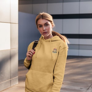 May include: A woman wearing a yellow hoodie with a mushroom graphic and the text "Shroom Queens". She is wearing blue jeans and has a black backpack slung over her shoulder.