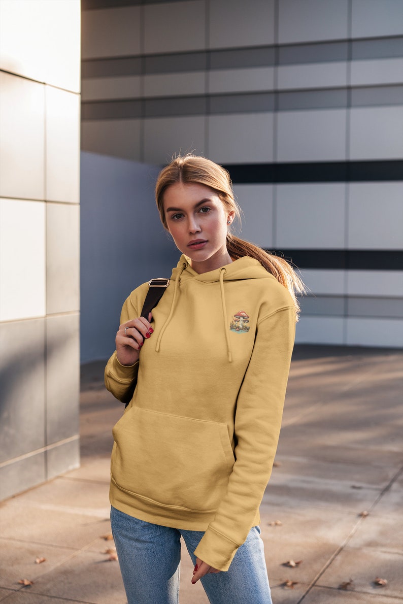 May include: A woman wearing a yellow hoodie with a mushroom graphic and the text "Shroom Queens". She is wearing blue jeans and has a black backpack slung over her shoulder.