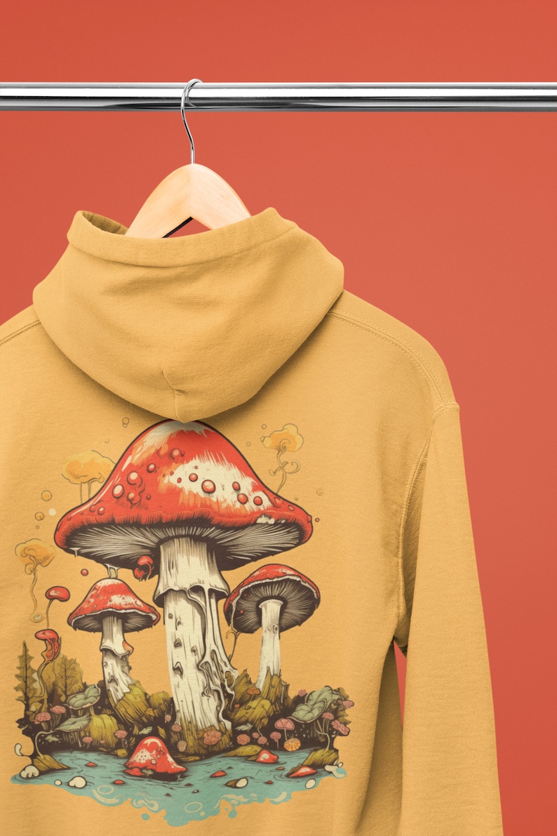 May include: A mustard yellow hoodie with a graphic of three red and white mushrooms in a forest setting. The mushrooms are surrounded by green foliage and a blue stream.
